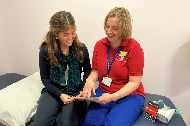 Two people sat down, smiling. A virtual reality headset is next to them. Laura, right, is wearing the red Play team t-shirt and is speaking with a teenage patient
