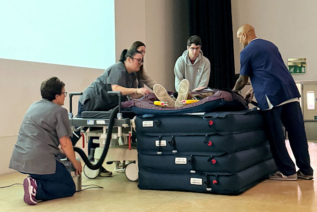 Five people on a stage lift a dummy on to a pile of stretchers