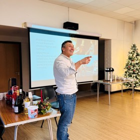A smiling male speaker in shirt and jeans; behind him a screen, Christmas tree, coffee table and table of Christmas gift items.