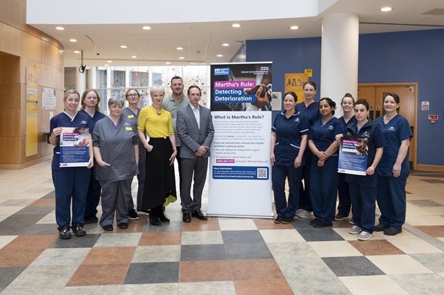 A group of healthcare staff standing in a hospital atrium beside a large “Martha’s Rule” information banner, with some team members holding promotional posters.
