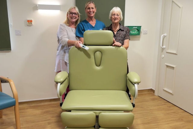 Three people behind a specialist green medical chair in a room with wooden flooring.