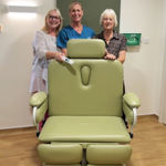 Three people behind a specialist green medical chair in a room with wooden flooring.