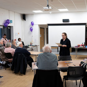 Person stood at front of room, holding a piece of paper, with other attendees watching