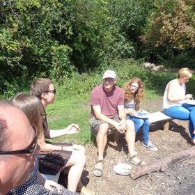A group of people outside, sat on benches and logs.
