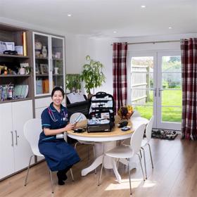A young woman in nurse's uniform sits at a circular table in a cheerful room with glass doors through which a sunny lawn is seen. An open toolbox of medical supplies is on the table.