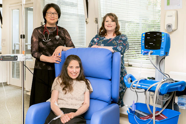 Young woman seated on padded chair in clinical setting, two older women in smart dresses stand behind