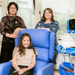 Young woman seated on padded chair in clinical setting, two older women in smart dresses stand behind