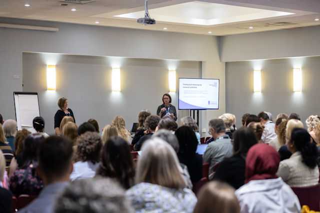 Two women with a screen and white board stand speaking to a large group of seated people