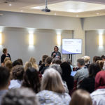 Two women with a screen and white board stand speaking to a large group of seated people