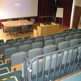 Looking down from upper level at back of small lecture theatre: six rows of seats, table at front with six chairs, large suspended screen behind.