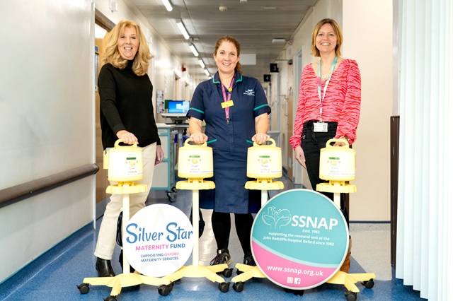 Three staff members in a hospital corridor standing behind four yellow breast pumps on wheeled stands, alongside display boards for the Silver Star Maternity Fund and SSNAP neonatal charity