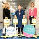 Three staff members in a hospital corridor standing behind four yellow breast pumps on wheeled stands, alongside display boards for the Silver Star Maternity Fund and SSNAP neonatal charity
