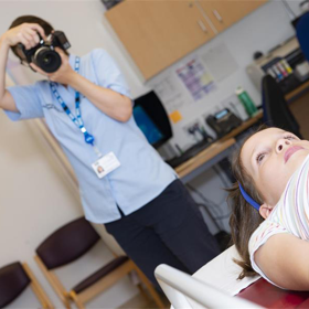 A photographer standing in a clinic room and pointing a camera at a young person lying down.