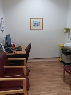 Consultation room with desk, computer, chairs, framed picture on wall and washbasin