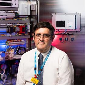 A man in a smart shirt and tie, lanyard and glasses, sits in front of a piece of complex machinery with multiple parts and wires.