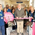 Group of nearly 20 people, some wearing clinical uniform. A patient in the middle prepares to cut a pink ribbon. There is a banner that reads "Oxford Hospitals Charity"