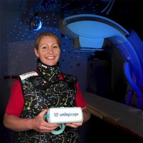 In a dark room a smiling woman in a sturdy vest-like garment holds a chunky headset in both hands. The arc of a large scanner is dimly illuminated alongside her.