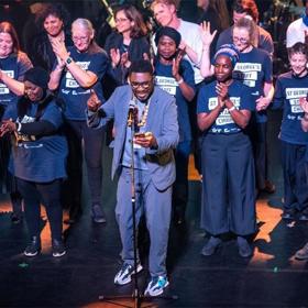 Around 20 applauding performers on stage in matching T-shirts; man in casual suit at front, hand raised, looking at mobile phone