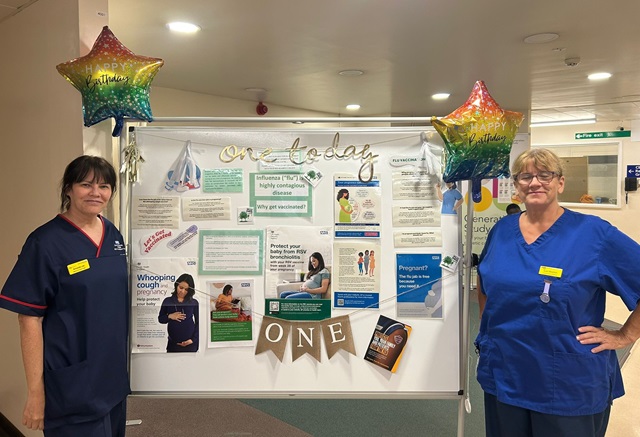 Two uniformed members of staff stand either side of a white board with information about vaccinations during pregnancy. Two balloons mark the first anniversary of the immunisation hubs