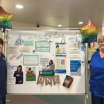 Two uniformed members of staff stand either side of a white board with information about vaccinations during pregnancy. Two balloons mark the first anniversary of the immunisation hubs