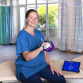A smiling woman in healthcare uniform sits on a bed in a therapy room holding a spherical object the size of a large grapefruit in her right hand. On the couch beside her rests a tablet computer with a diagram on the screen.