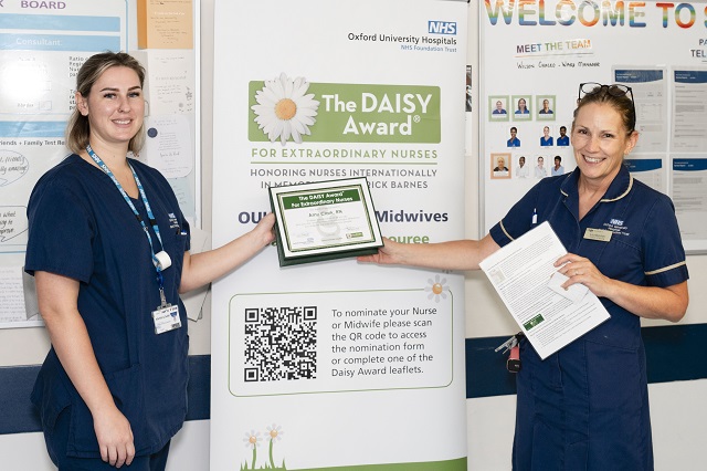 Two staff members at OUH stand in front of a DAISY Award display banner. One holds a framed certificate, the other a nomination leaflet. The banner promotes the DAISY Award for Extraordinary Nurses and Midwives, featuring a QR code for nominations.