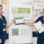 Two staff members at OUH stand in front of a DAISY Award display banner. One holds a framed certificate, the other a nomination leaflet. The banner promotes the DAISY Award for Extraordinary Nurses and Midwives, featuring a QR code for nominations.
