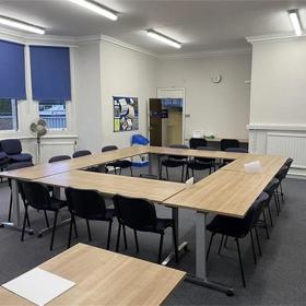 Smooth top desk alongside four tables arranged in a square, four chairs each side: open door behind, strip lights above.